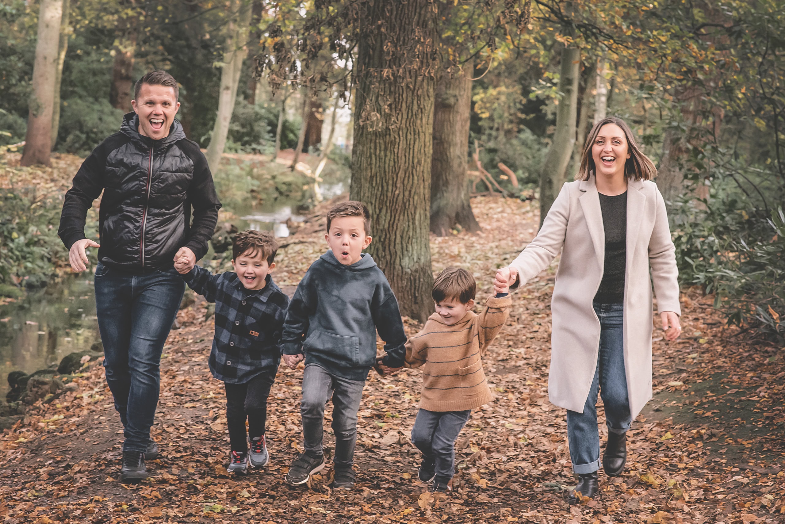Family of five running hand in hand through autumn leaves in the woods, all laughing