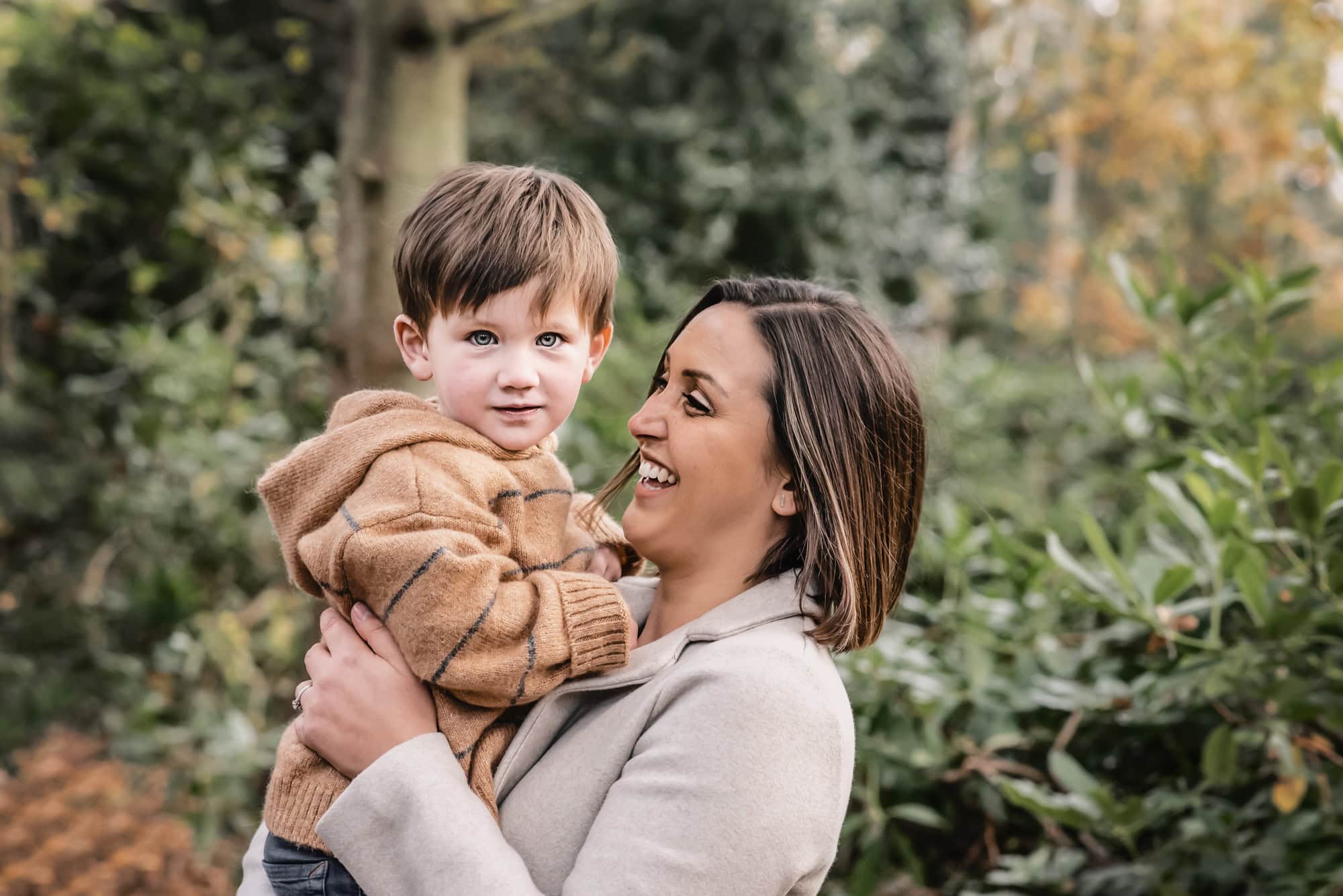 Mum laughing as she holds her toddler son in a striped jumper among autumn trees