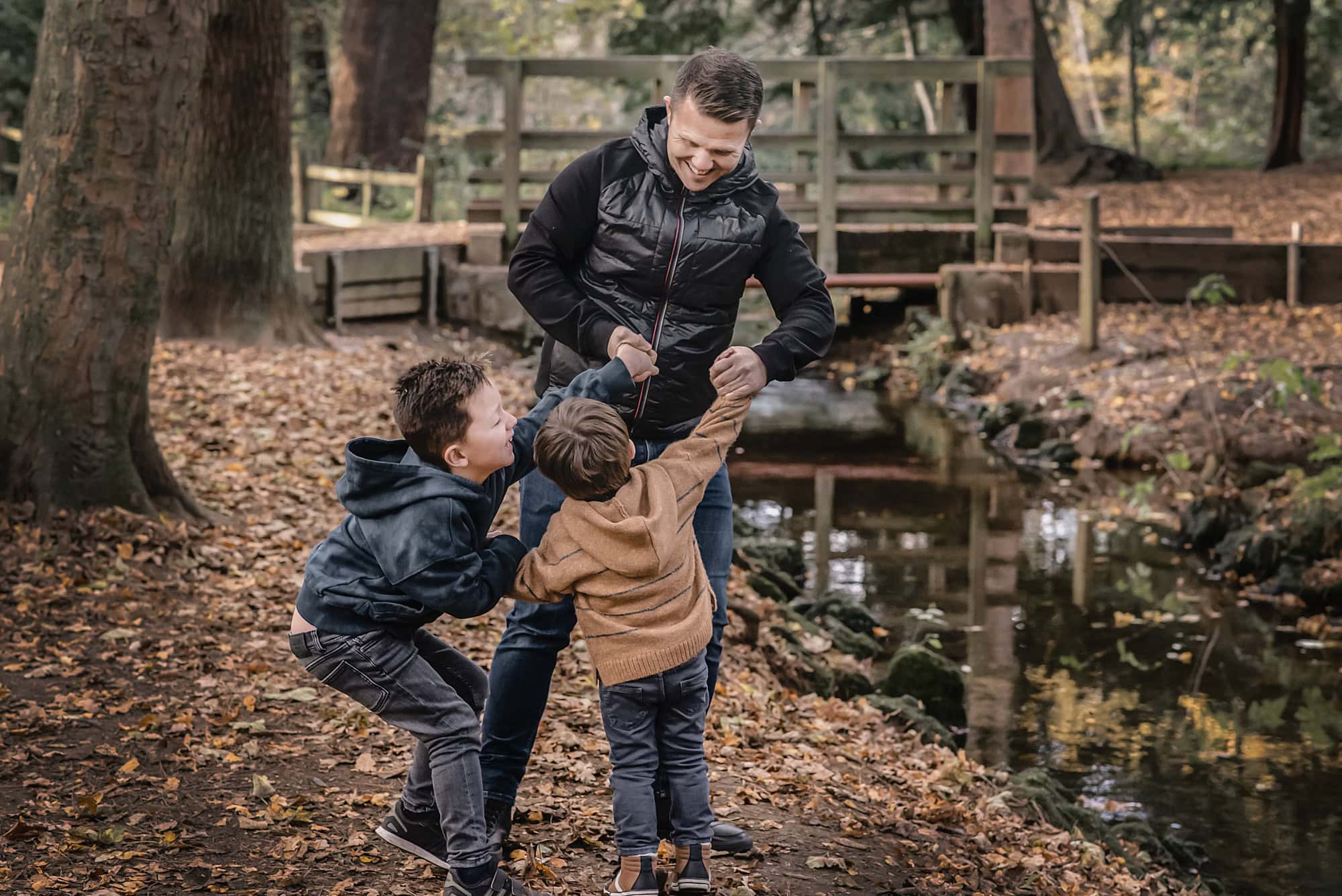 Dad laughing as his two sons pull on his arms beside a woodland stream in autumn