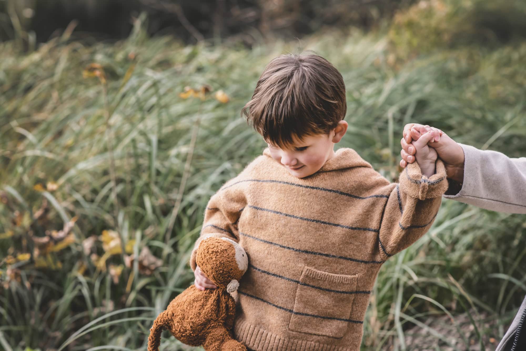 Little boy in a striped knitted hoodie holding his toy monkey and his mum's hand on an autumn walk